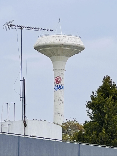 上部が円盤のようになっている、白い塔があり、円盤の下の方には赤く丸い宇宙開発事業団のロゴと、青いJAXAのロゴが描かれています